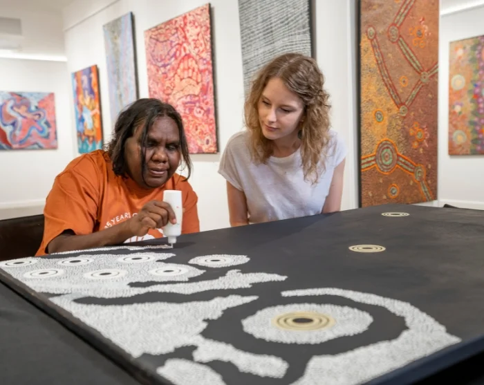 Taiteilija maalaa aboriginaalitaidetta Australian Gallery of Central Australia -galleriassa naisen katsellessa vieressä