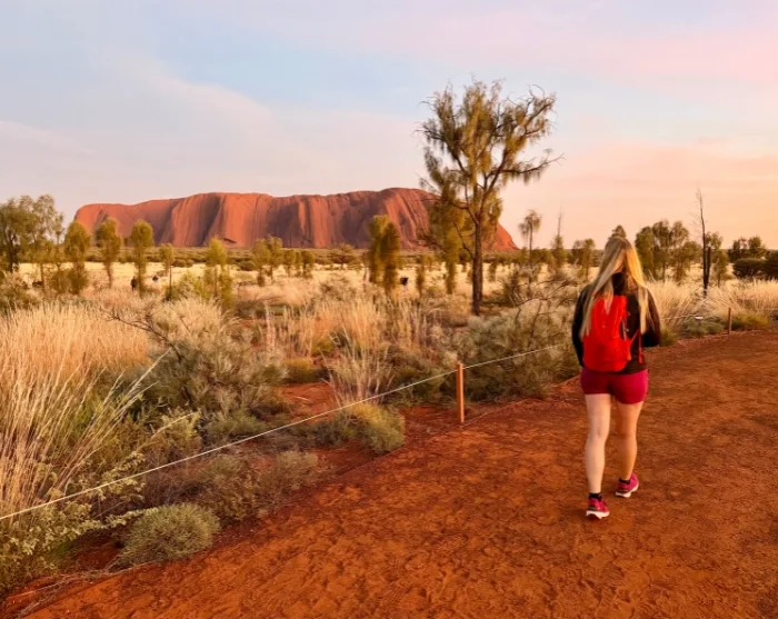 Nainen kulkee polkua pitkin auringon noustessa Uluru-kallion taustalla Uluru–Kata Tjutan kansallispuistossa Australiassa