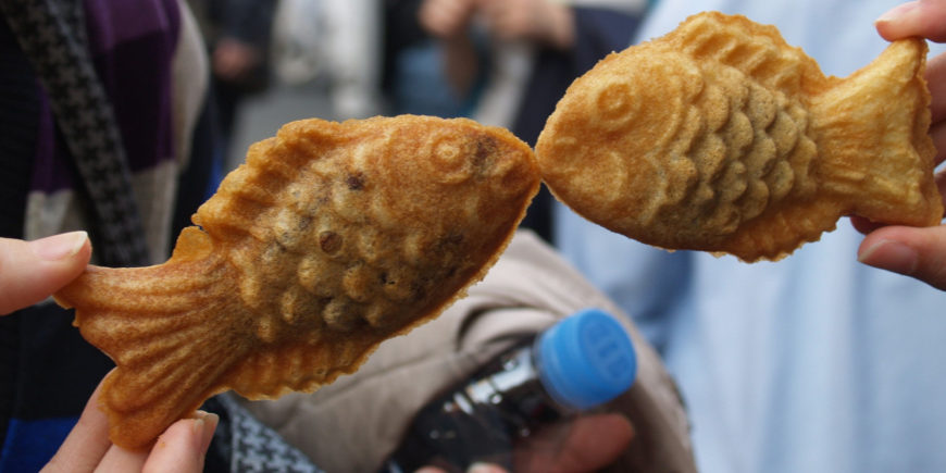 Kaksi henkilöä pitelee taiyaki-leivonnaisia niin, että ne näyttävät suutelevilta kaloilta